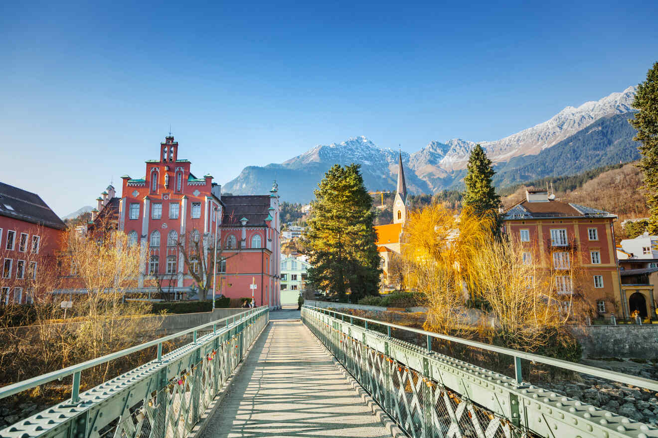 Historic red building in Innsbruck beside a river, with a bridge leading towards it and the Alps in the distance